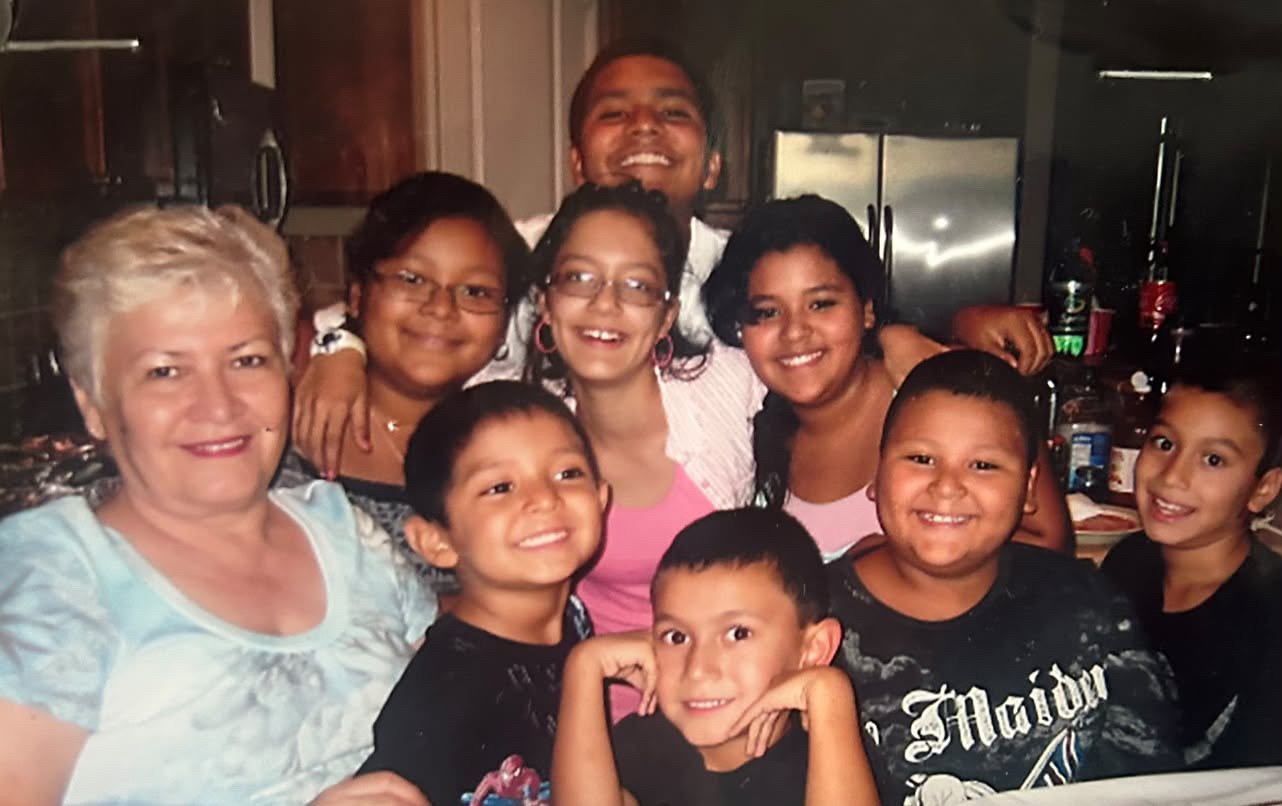 A smiling grandmother surrounded by seven grandchildren gathered closely in a kitchen