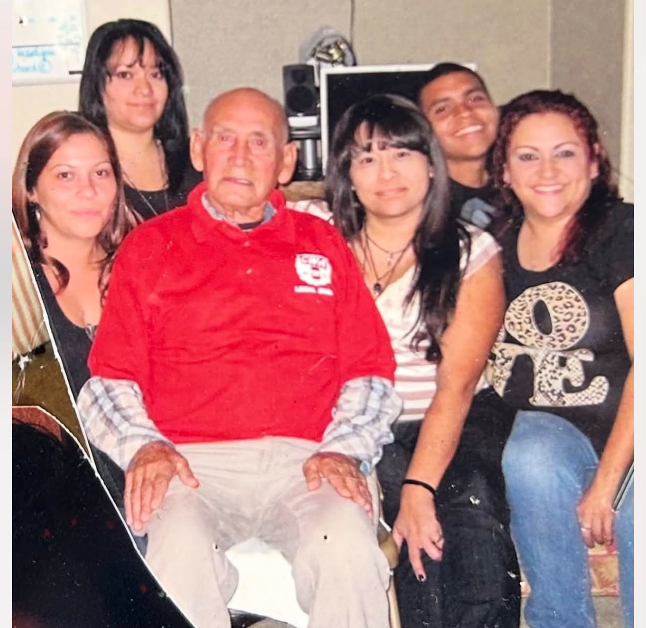 An elderly man in a red polo shirt with five younger adults gathered close around him indoors